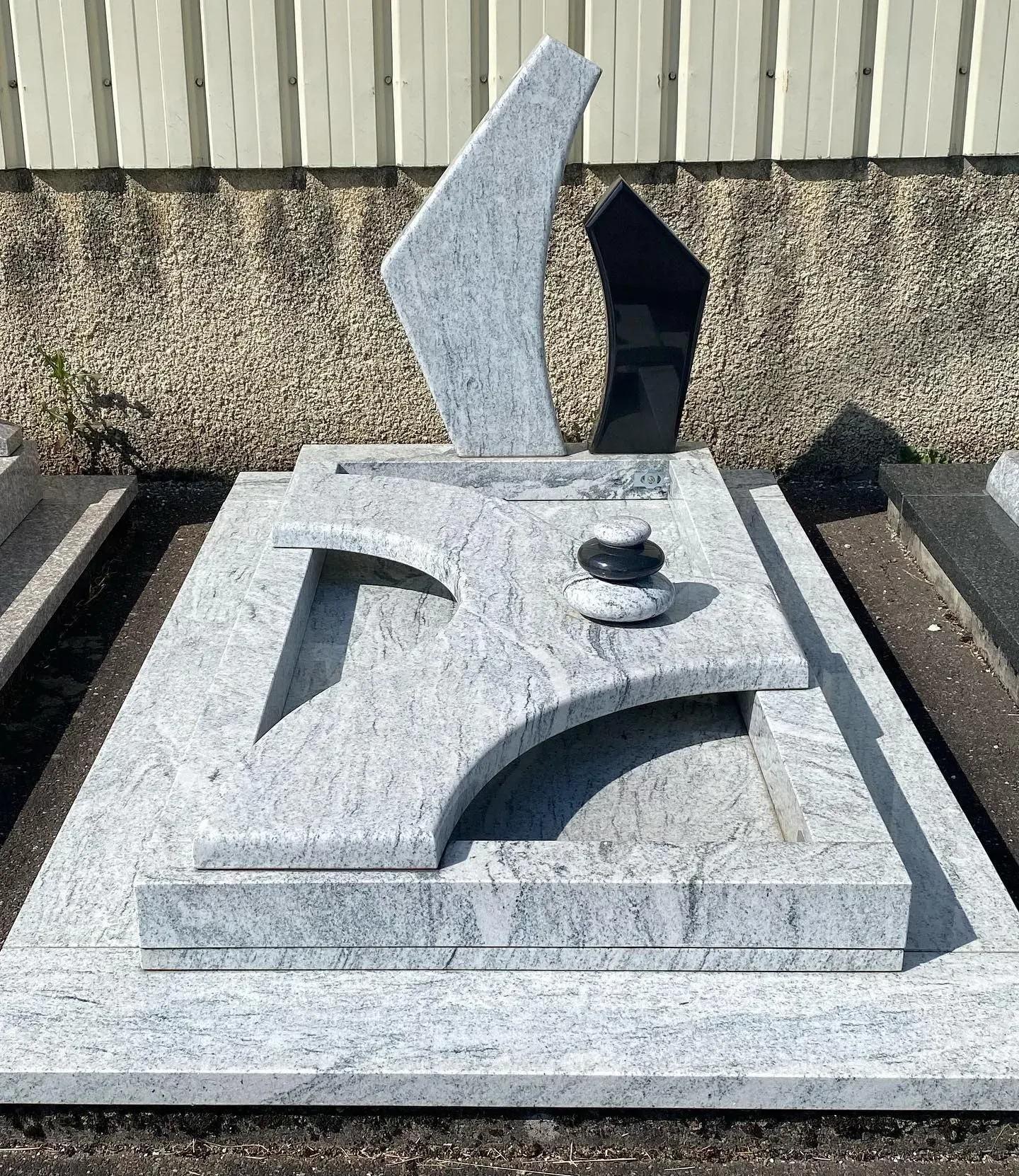 Monument funéraire moderne en granit clair veiné avec éléments gravés et galets décoratifs.